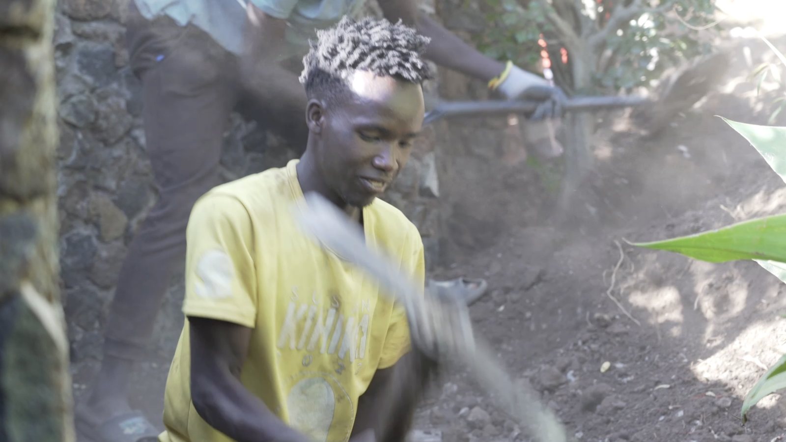 A young apprentice mid-swing with a pickaxe, dust in the air, dry-stone wall rising behind